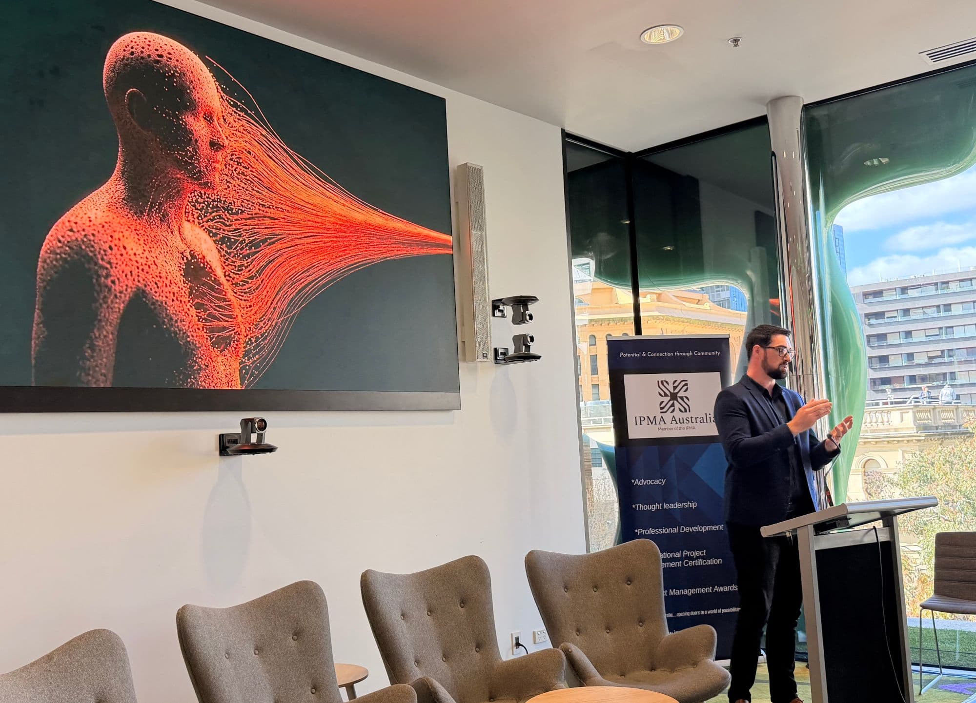 Josh Horneman delivering the Curiosity Agent keynote for IPMA Australia, beside a large screen showing a human figure dissolving into streams of orange light